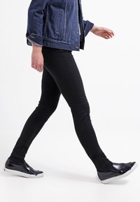 Jeans skinny negros combinados con una chaqueta de mezclilla azul y zapatos negros sin cordones con suela blanca. El conjunto presenta un diseño casual y ajustado.