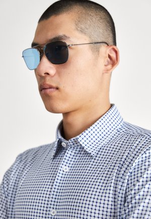Chemise à manches courtes à carreaux blanche et marine avec un col boutonné. Le modèle porte des lunettes de soleil carrées teintées en noir et en clair.