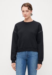 Felpa nera con dettagli di borchie sulle spalle, scollatura a girocollo e polsini a coste, abbinata a jeans blu. Tessuto morbido e vestibilità comoda.