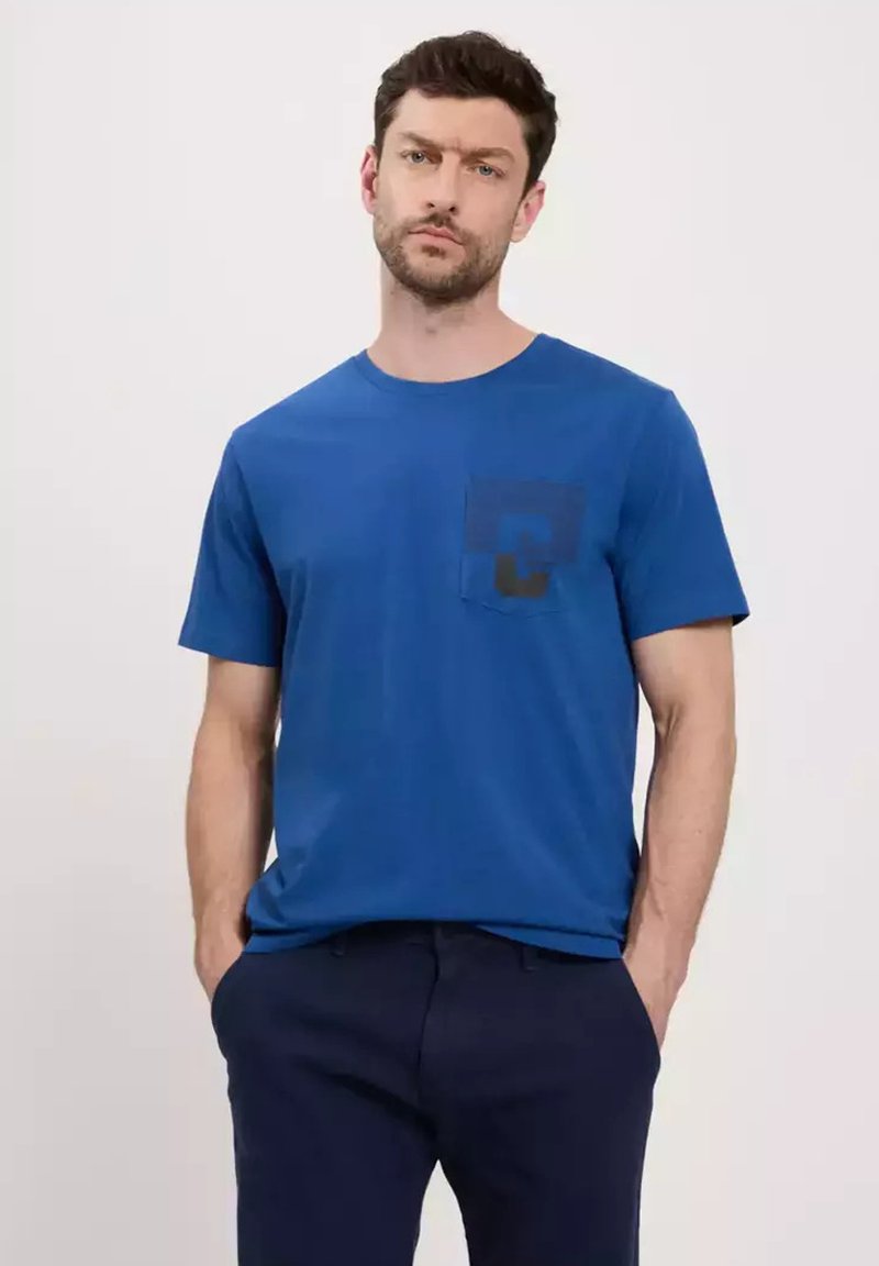 Niebieska bawełniana koszulka z okrągłym dekoltem i krótkimi rękawami. Posiada czarną kieszeń z geometrycznym wzorem. Noszona z ciemnymi spodniami.