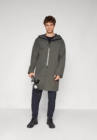 Vodootporni maslinasto zeleni kaput s prednjim zatvaračem, kapuljačom i dugim rukavima, uparen s tamnim hlačama i crnim cipelama, držeći bučice.