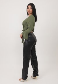Top avvolgente a maniche lunghe color verde oliva con maniche a campana abbinato a pantaloni in pelle neri a vita alta. Tacchi bianchi aperti sulla punta visibili.