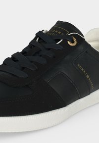 Μαύρο δερμάτινο αθλητικό παπούτσι Tommy Hilfiger με μαύρα κορδόνια, χρυσό οφθαλμό και λευκή σόλα, με το όνομα της μάρκας στο πλάι και στη γλώσσα.