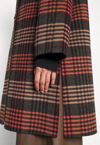 Mano descansando al lado, vistiendo un abrigo a cuadros en marrón, rojo y beige, con una manga negra debajo, contra un fondo sencillo.