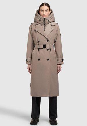 Trench-coat marron clair avec une grande capuche, boutons double boutonnage, détail de ceinture et manches longues ; porté par-dessus un pantalon noir et des bottes.