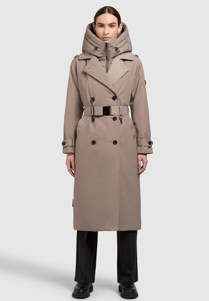 Lichtbruine trenchcoat met een grote capuchon, dubbelrijgsluitingen, riemdetail en lange mouwen; gedragen over zwarte broek en laarzen.
