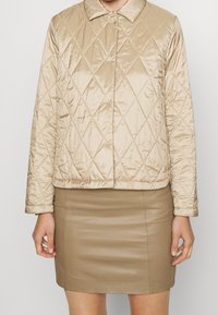 Beige quilted jacket with a collar, featuring a shiny texture and diamond pattern. Worn over a fitted beige leather skirt.