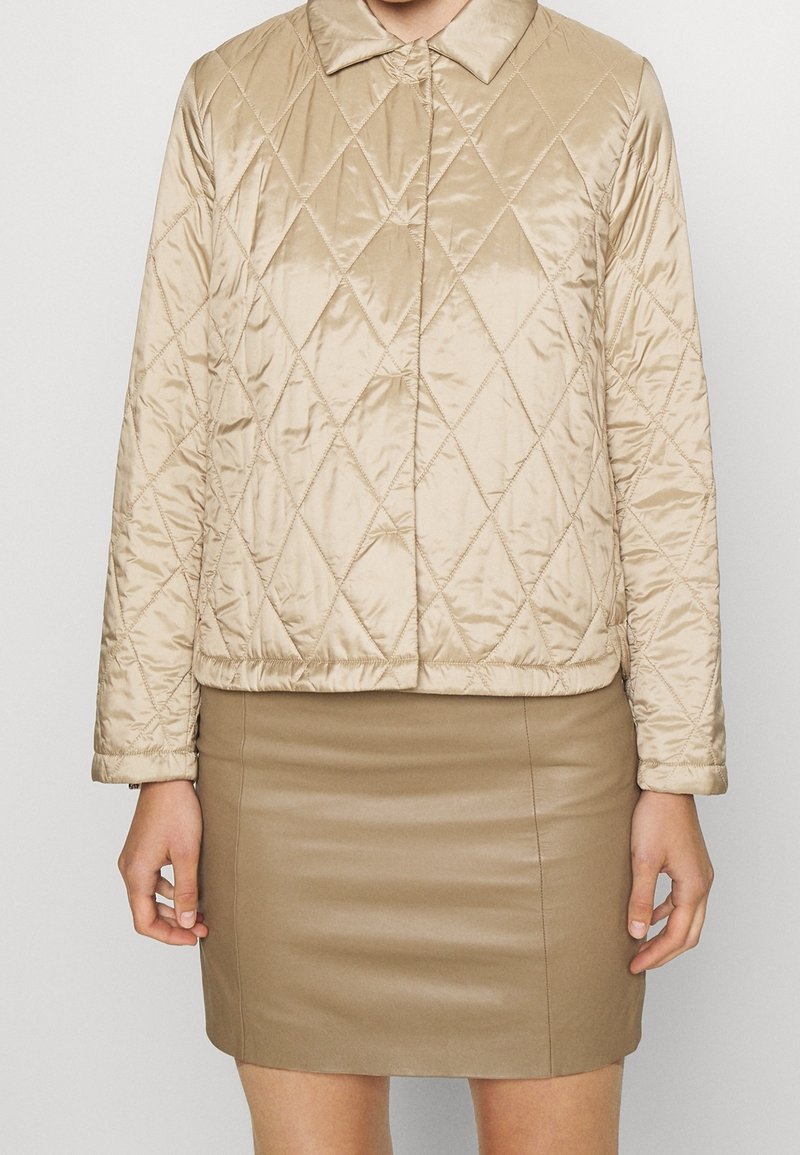 Beige quilted jacket with a collar, featuring a shiny texture and diamond pattern. Worn over a fitted beige leather skirt.