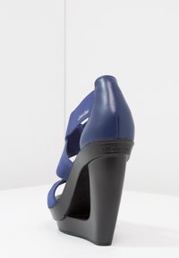 Sandale à talon compensé bleu avec une tige en tissu texturé et des sangles élastiques, dotée d'une plateforme noire élégante. Logo Calvin Klein sur le talon.