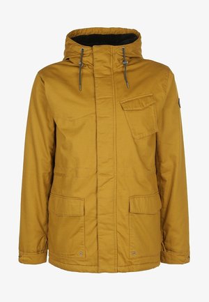 Homme portant une veste jaune moutarde, un pull en maille torsadée blanc, un pantalon de neige noir et des chaussures de randonnée noires, debout les mains dans les poches.