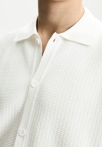 Weißes, strukturiertes, gestricktes Polohemd mit einem Kragen, ausgestattet mit einer Knopfleiste und dezenten horizontalen Streifen. Die Knöpfe sind rund und weiß.