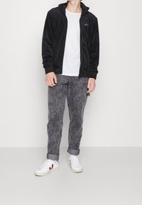 Chaqueta negra de forro polar con cremallera y cuello alto, combinada con jeans grises texturizados y zapatillas blancas con detalles en rojo.