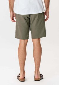 Mand iført olivengrønne knælange shorts, en hvid skjorte og mørkebrune slip-on sandaler, står med ryggen til på en hvid baggrund.