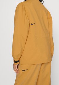 Nike-jacka i senapsgul, tillverkad av mjukt tyg, med puffärmar, svarta detaljer och en liten logotyp på baksidan.