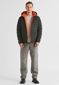 Mann steht gerade und trägt eine schwarze Jacke mit orangefarbener Innenausstattung, ein beiges Hemd, ausgebleichte graue Jeans und schwarze Stiefel vor einfarbigem Hintergrund.