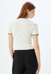 Weißes, geripptes Kurzarmoberteil mit schwarzem Besatz, figurbetontes Design, strukturiertes Gewebe, getragen mit schwarzen Hosen. Rückansicht zeigt eine strukturierte Silhouette.