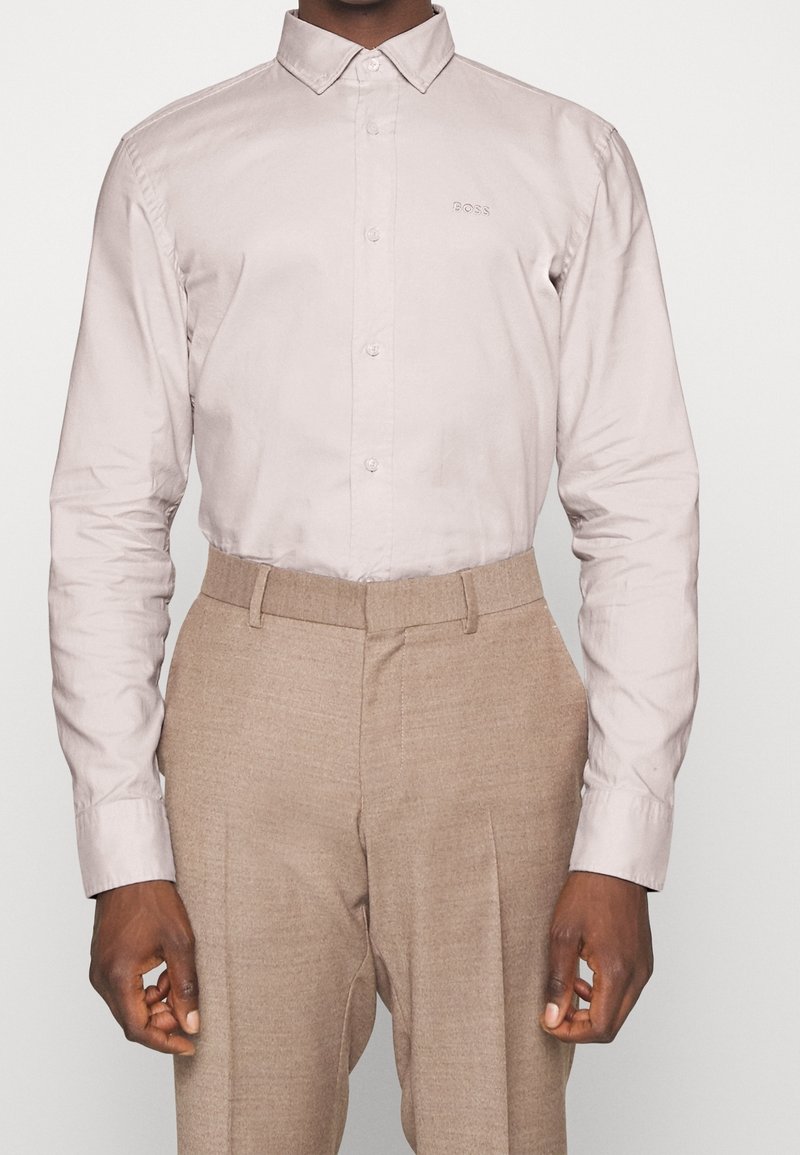 Homme portant une chemise beige clair boutonnée rentrée dans un pantalon en laine beige à taille haute, sur un fond uni.