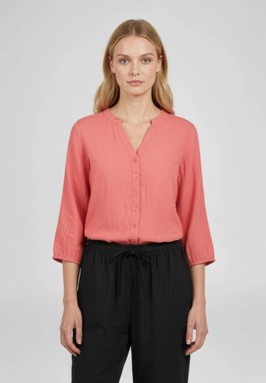 Frau mit heller Haut und blondem Haar, die eine korallenfarbene Bluse mit Knöpfen und 3/4-Ärmeln sowie schwarze Hose mit Kordelzug trägt, blickt frontal vor einem grauen Hintergrund.