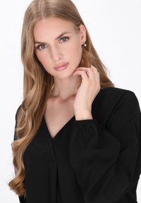 Blusa nera con scollatura a V e maniche lunghe leggermente a sbuffo. Tessuto morbido con un leggero luccichio. Il modello ha lunghi capelli mossi e un orecchino delicato.