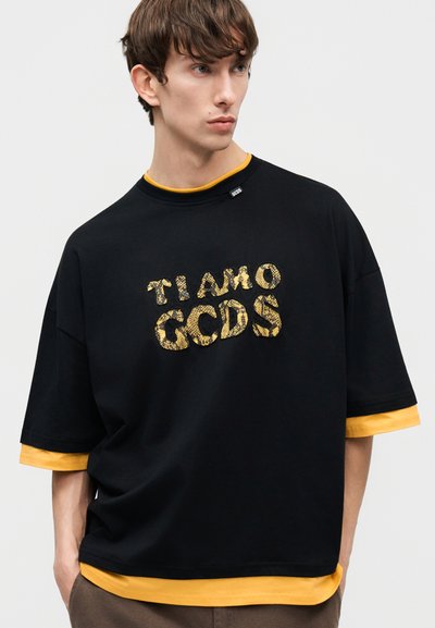 Junger Mann trägt ein übergroßes schwarzes T-Shirt mit gelbem Schlangenmuster-Text "TIAMO GCDS", darüber eine gelbe Langarmshirt.