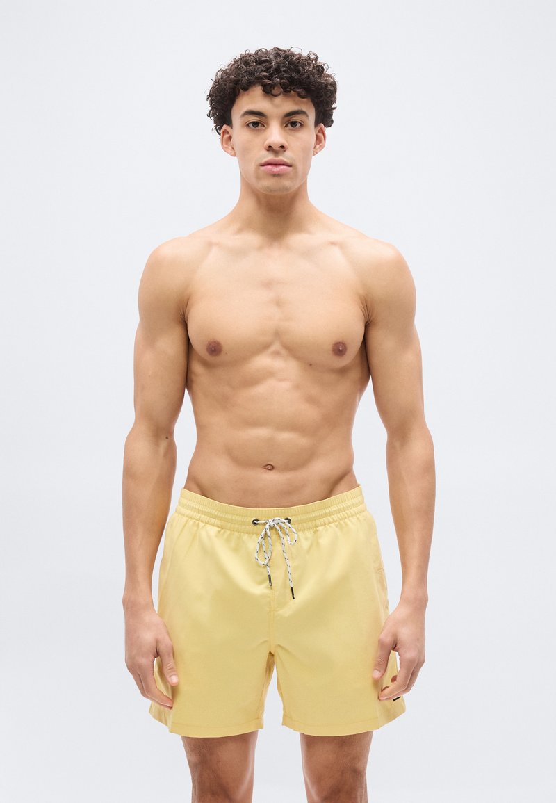 Νεαρός μυώδης άνδρας που στέκεται και κοιτάζει μπροστά, φορώντας κίτρινο μαγιό με κορδόνι, πάνω σε απλό φωτεινό φόντο