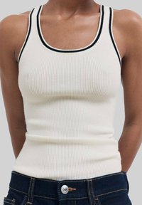 Gerippter, cremefarbener Tanktop mit schwarzem Besatz am Halsausschnitt und an den Armlöchern; taillierte Form; getragen mit dunklen Jeans.