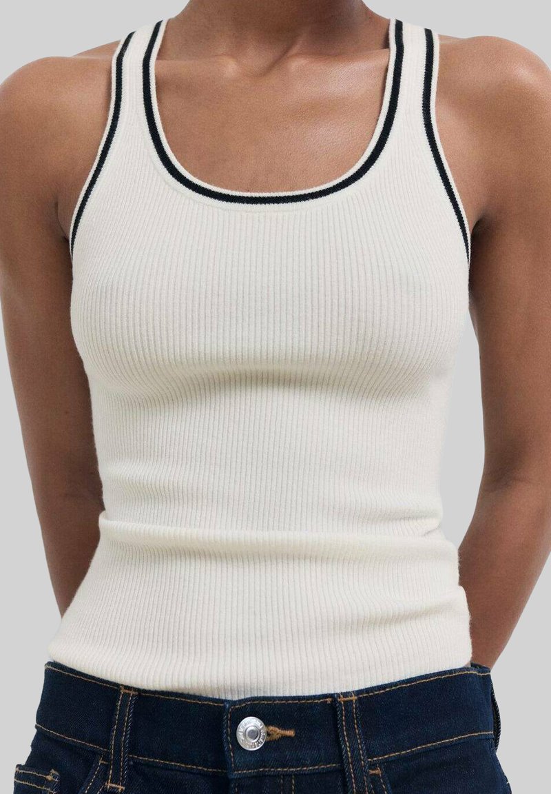 Gerippter, cremefarbener Tanktop mit schwarzem Besatz am Halsausschnitt und an den Armlöchern; taillierte Form; getragen mit dunklen Jeans.