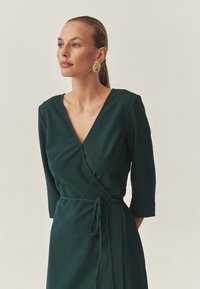 Robe verte émeraude, de style enveloppant, avec des manches trois-quarts et une taille nouée. Texture lisse avec un décolleté en V profond et des détails de couture subtils.