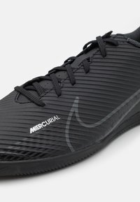 Musta urheilukenkä, jossa on tekstuuripintainen päällinen, joka sisältää vaakasuoria uria, nauhat ja hillityn Nike-logon sivussa. Kuminpohja pitoa varten.