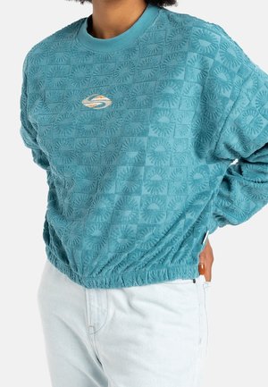 Personne portant un sweat-shirt texturé teal avec des carrés à motif soleil et un logo sur la poitrine, associé à un jean bleu clair, main sur la hanche.