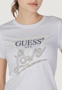 Witte katoenen T-shirt met een zwart "GUESS U.S.A." logo en strassversiering die "Love" spelt, met daarachter een driehoekgrafiek.