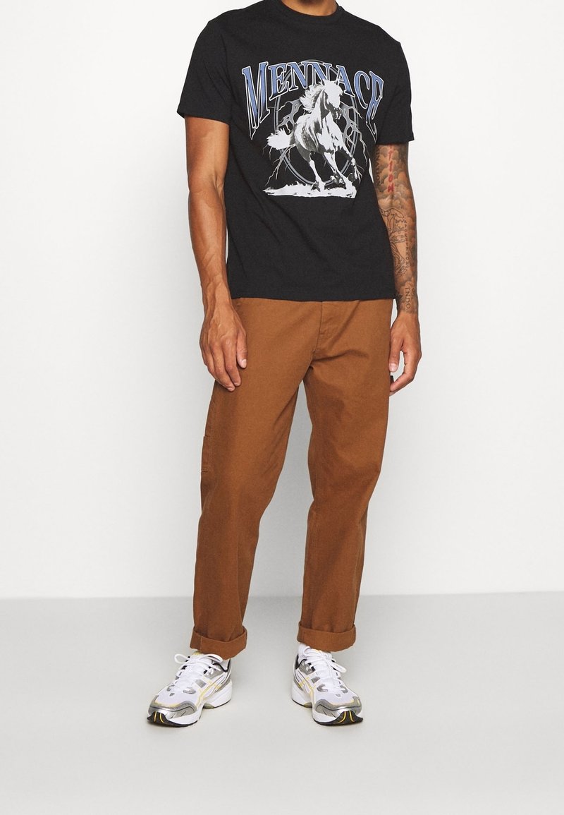 T-shirt graphique noir avec un design de cheval, associé à un pantalon marron et des baskets blanches avec des accents argentés et dorés.