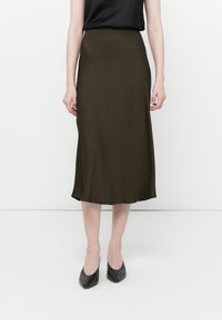 Jupe midi vert olive en tissu lisse avec un léger éclat, dotée d'une taille cintrée et d'une silhouette évasée, accompagnée de talons noirs.