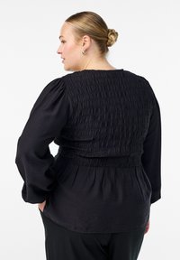 Naine, kellel on juuksed tõstetud kuklasse, kannab hõbedaseid kõrvarõngaid ja musta pikakäepluusiga, millel on tekstureeritud smockdetail tagaküljel, vaadates tagant.