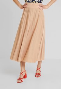 Gonna midi plissettata in tessuto beige chiaro con una cintura larga e una caduta fluida, abbinata a sandali con tacco a strisce rossi.