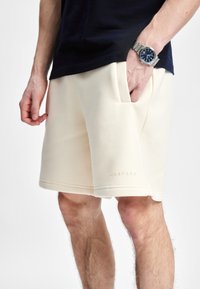Kremfargede shorts laget av myk, teksturert stoff, med sidelommer og en sømt kant. Merkenavnet "CASPARA" er diskret preget.