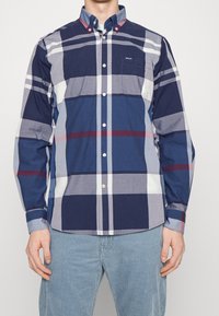 Geruit overhemd met knopen in marineblauw, grijs en wit met rode accenten. Katoenen stof, lange mouwen en een borstzakje.