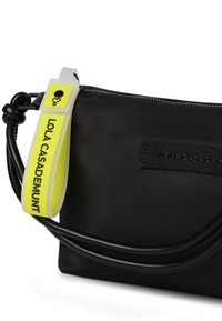 Czarna torba z materiału z zamkiem błyskawicznym, z żółtą plastikową etykietą z napisem "LOLA CASADEMUNT" oraz czarnym skórzanym paskiem o gładkiej fakturze.