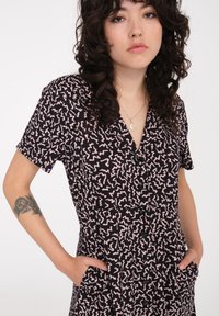 Schwarzes Kleid mit pinkem abstraktem Muster, kurzen Ärmeln, V-Ausschnitt, Knopfverschluss vorne und Seitentaschen. Glattes Material mit subtiler Textur.