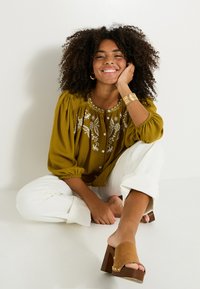 Blouse vert olive avec broderie florale, devant à boutons et manches bouffantes, associée à un pantalon blanc à jambes larges et des mules à talons bruns.