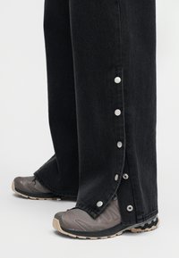 Fekete bőszárú nadrág ezüst patentgombokkal a külső oldalszegély mentén, szürke és bézs túracipők fölött viselve.