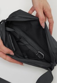 Borsa a tracolla nera con chiusura a zip, che mostra i compartimenti interni, le cinghie e un'etichetta che recita "Fatta di plastica ad impatto oceano". Tessuto liscio.