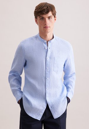 Junger Mann trägt ein hellblaues langärmeliges Hemd mit Stehkragen und Knöpfen, Hände in den Taschen, vor einem schlichten hellen Hintergrund.
