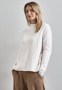 Maglione bianco lavorato a maglia con design a testa di montone, colletto e orlo a costine, texture morbida, indossato con pantaloni marroni, dal taglio morbido.