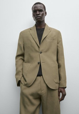 Olijfgroen linnen pak met een gestructureerde blazer, voorzien van twee knopen, notch revers en opgestikte zakken. Draagt over een zwart shirt.