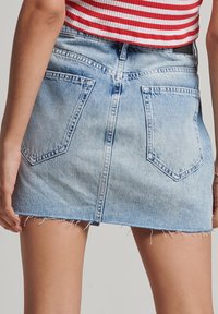 Leren rok in lichtblauw met een rafelige onderrand, twee achterzakken en een centrale naad. Materiaal is gestructureerde denim met vervaagde gebieden.