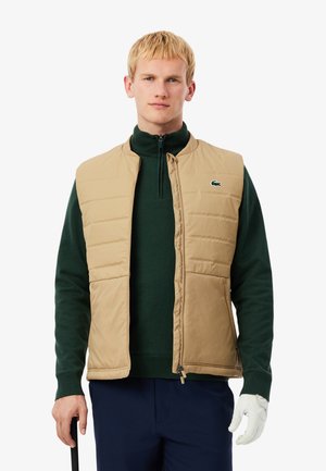 Veste rembourrée beige avec col montant, portée sur un pull vert foncé. Dotée d'une fermeture éclair et de poches latérales. Logo Lacoste sur la poitrine.