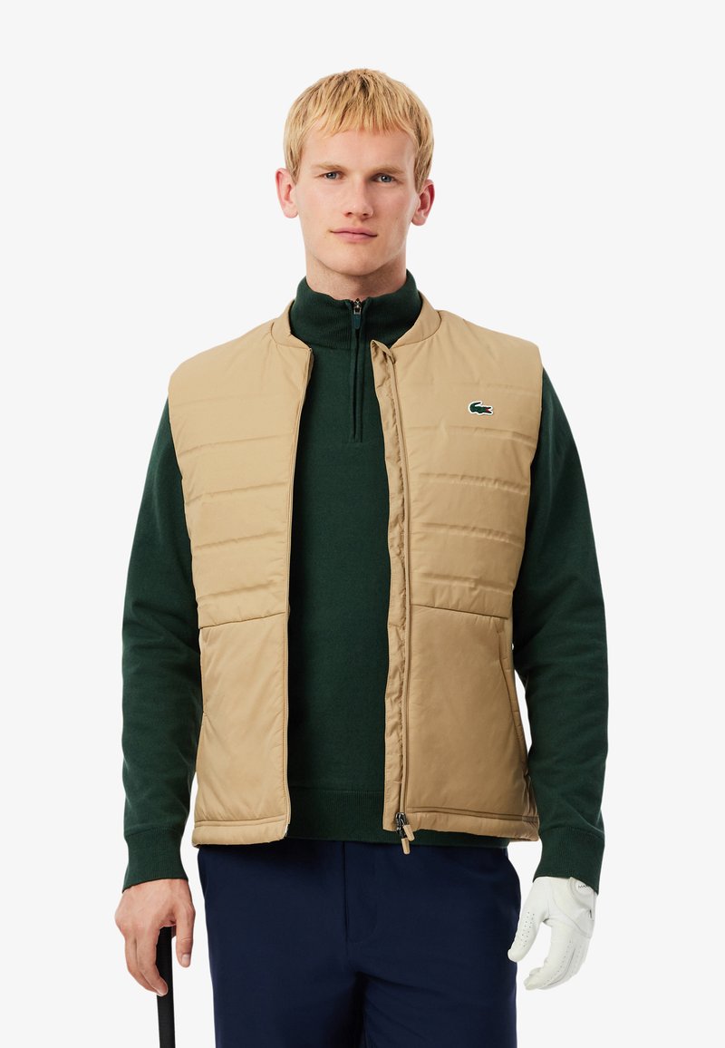 Chaleco acolchado beige con cuello alto, llevado sobre un suéter verde oscuro. Cuenta con cierre de cremallera y bolsillos laterales. Logotipo de Lacoste en el pecho.