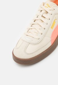 Beige sportcipő velúr és bőr anyagból, kerek orral, texturált barna talppal és élénk narancs kiegészítőkkel. Fűzős kialakítással rendelkezik.