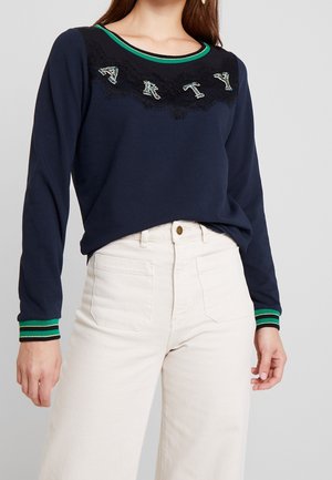 Femme portant un haut à manches longues bleu marine avec des poignets et un col rayés verts, un pantalon blanc taille haute, et "ARTY" orné sur la poitrine.